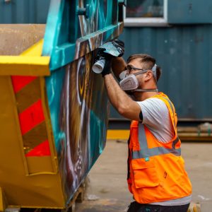 Artist spray painting a skip with vibrant colours as part of the Skip Art Challenge, transforming it into a creative and eye-catching piece of artwork