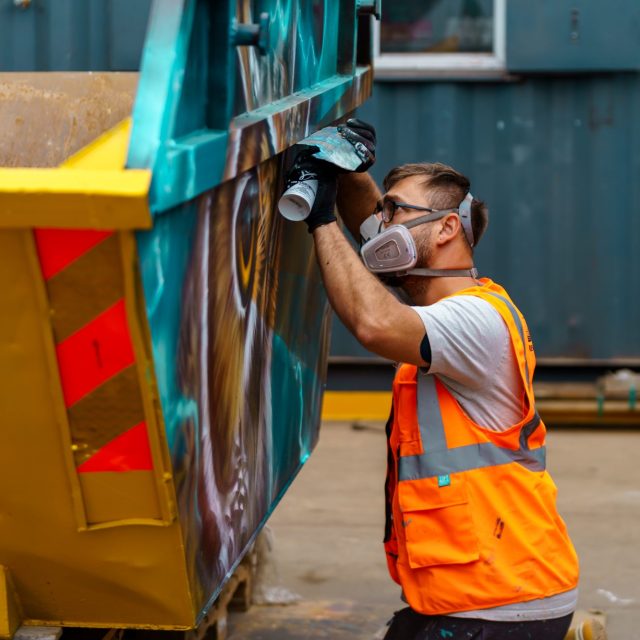 Artist spray painting a skip with vibrant colours as part of the Skip Art Challenge, transforming it into a creative and eye-catching piece of artwork
