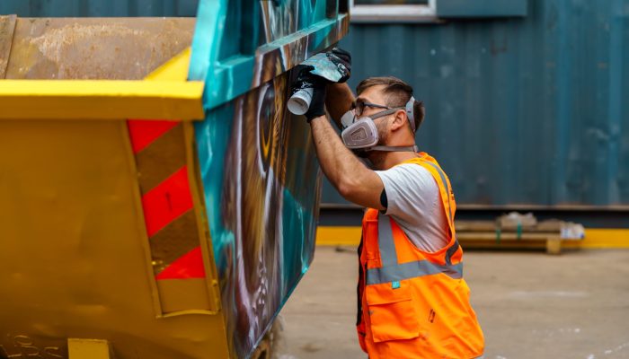 Artist spray painting a skip with vibrant colours as part of the Skip Art Challenge, transforming it into a creative and eye-catching piece of artwork